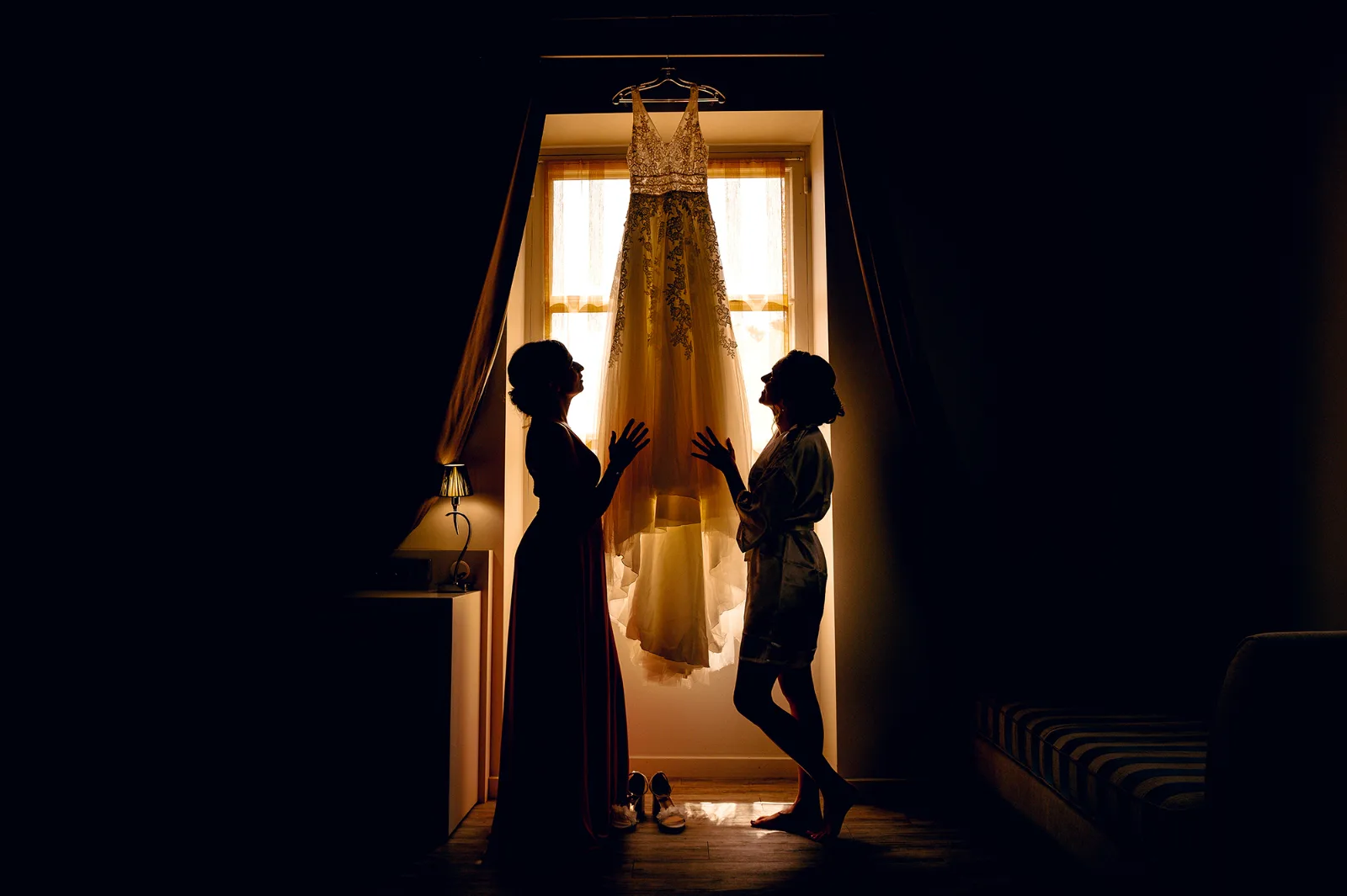 Robe de mariée suspendue devant une fenêtre, silhouettes de deux femmes, préparatifs de la mariée